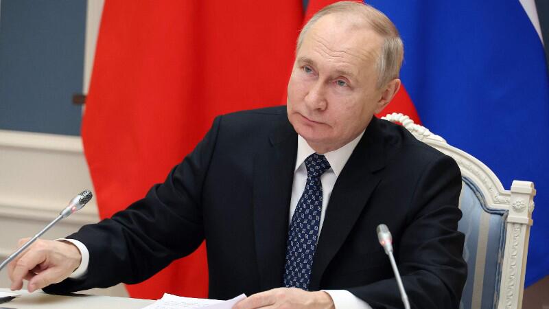 Wladimir Putin sitzt am Tisch vor einer Russland-Fahne.