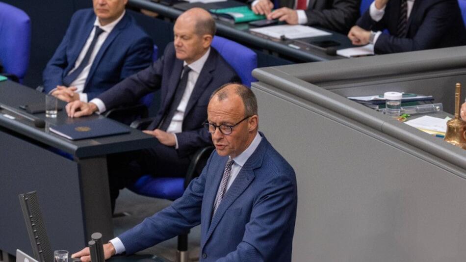 Olaf Scholz sitzt im Bundestag hinter Friedrich Merz, wenn dieser eine Rede hält.