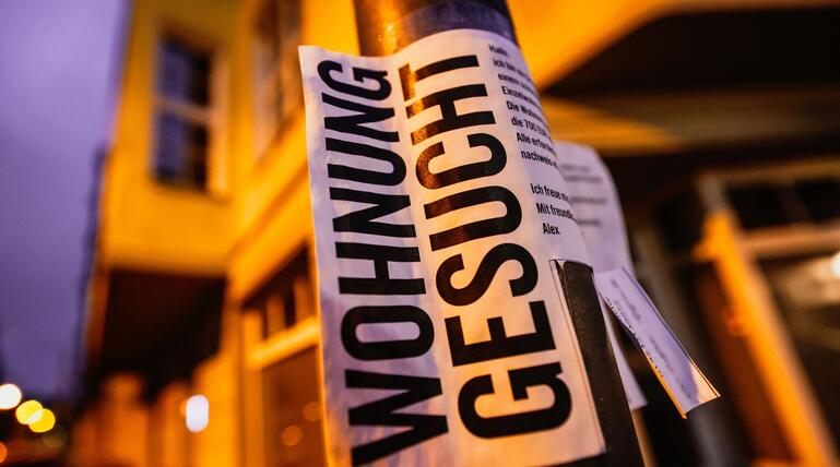 Ein Zettel mit der Aufschrift "Wohnung gesucht" vor Wohnhäusern