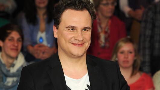 Guido Maria Kretschmer lächelt bei Talkshow. Er trägt einen schwarzem Blazer und ein weißes Shirt.
